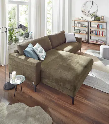 Großes grünes Ecksofa in modernem Wohnzimmer mit Holzfußboden, hellen Vorhängen und dekorativen Kissen. Helles Tageslicht, Pflanzen und Bücherregal schaffen eine gemütliche Wohnatmosphäre.